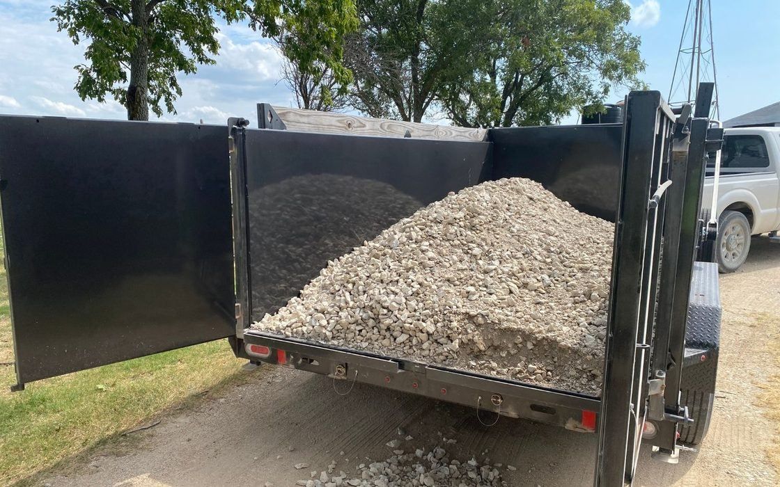 A dump trailer loaded with crushed stone, tailgate open, on a gravel drive